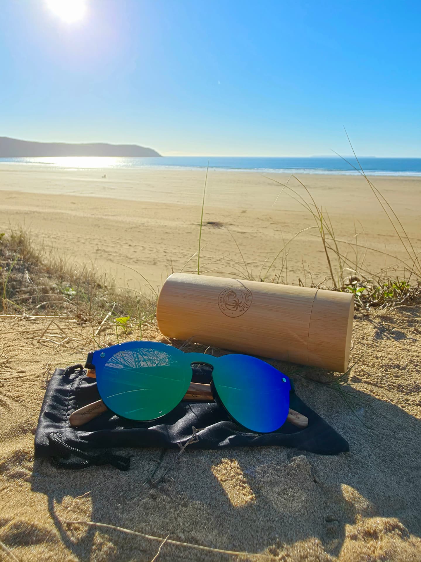 Tide & Palm sunglasses resting on the black pouch with the engraved bamboo case behind, on the beach above the sea